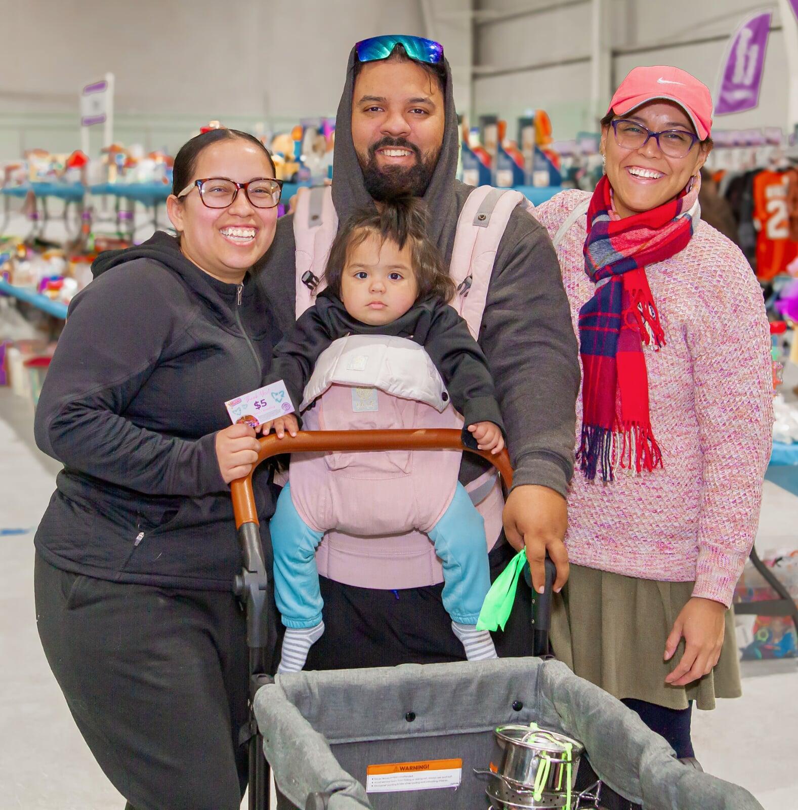 A mother carries her son in a front carrier. Grandmother is pushing the stroller. They are all shopping at the JBF sale and are surrounded by toys.