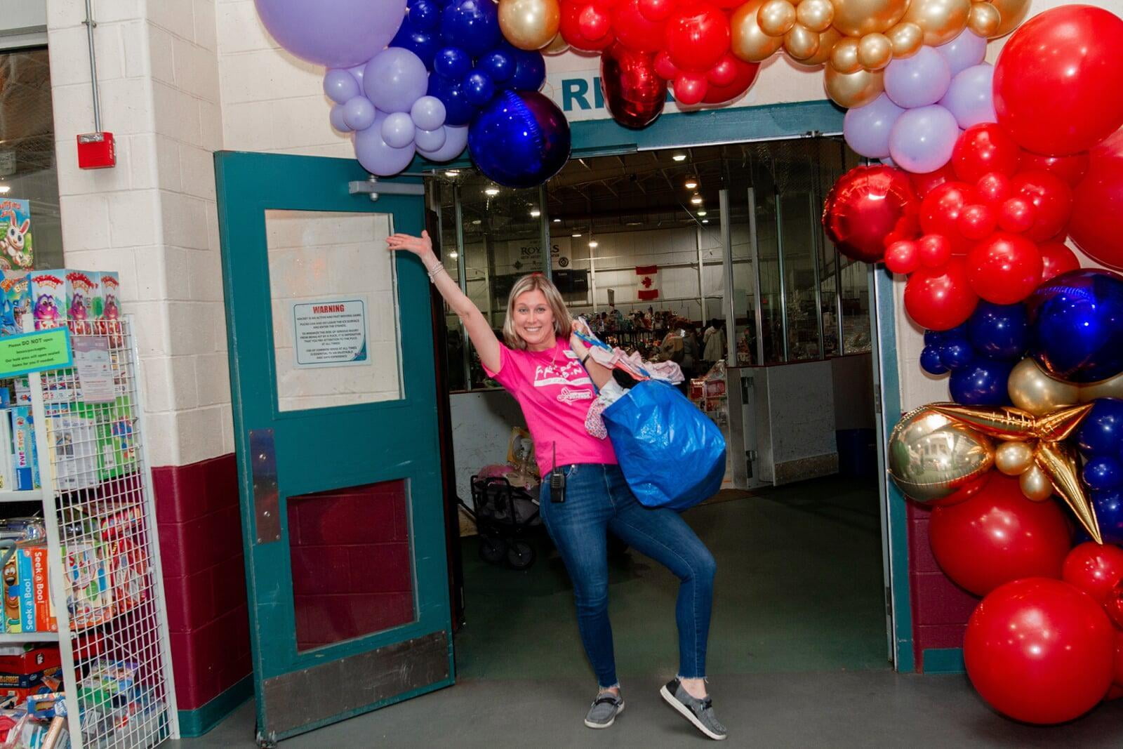 Woman waving inside of a doorway. The doorway is surrounded in purple, blue, gold and red balloons.