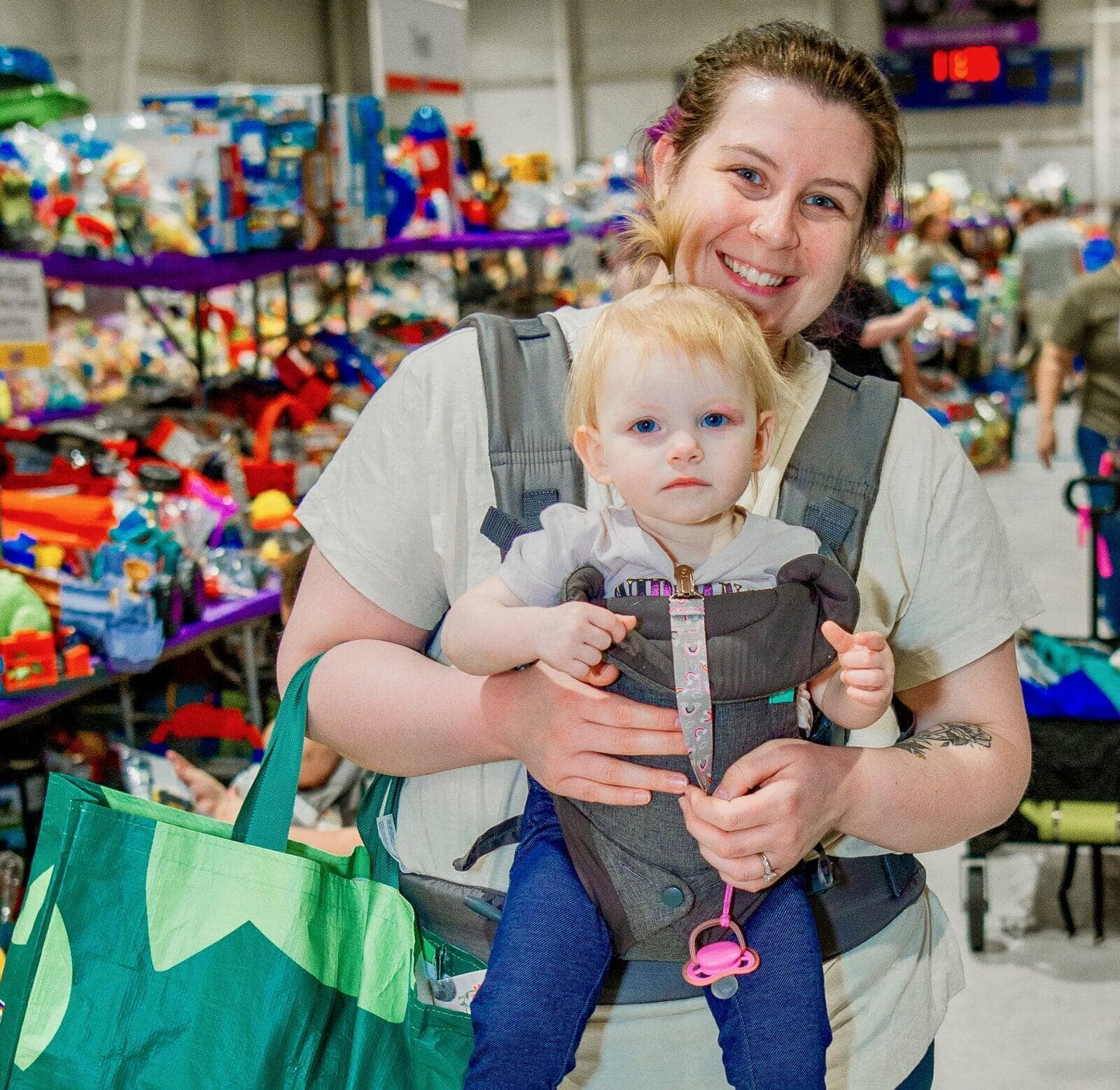 Woman carrying toddler in front carrier smiling.  Tables full of toys are in the background.