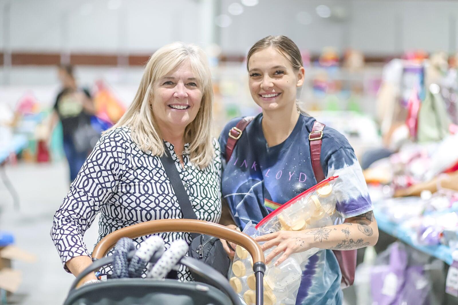 A mother carries her son in a front carrier. Grandmother is pushing the stroller. They are all shopping at the JBF sale and are surrounded by toys.