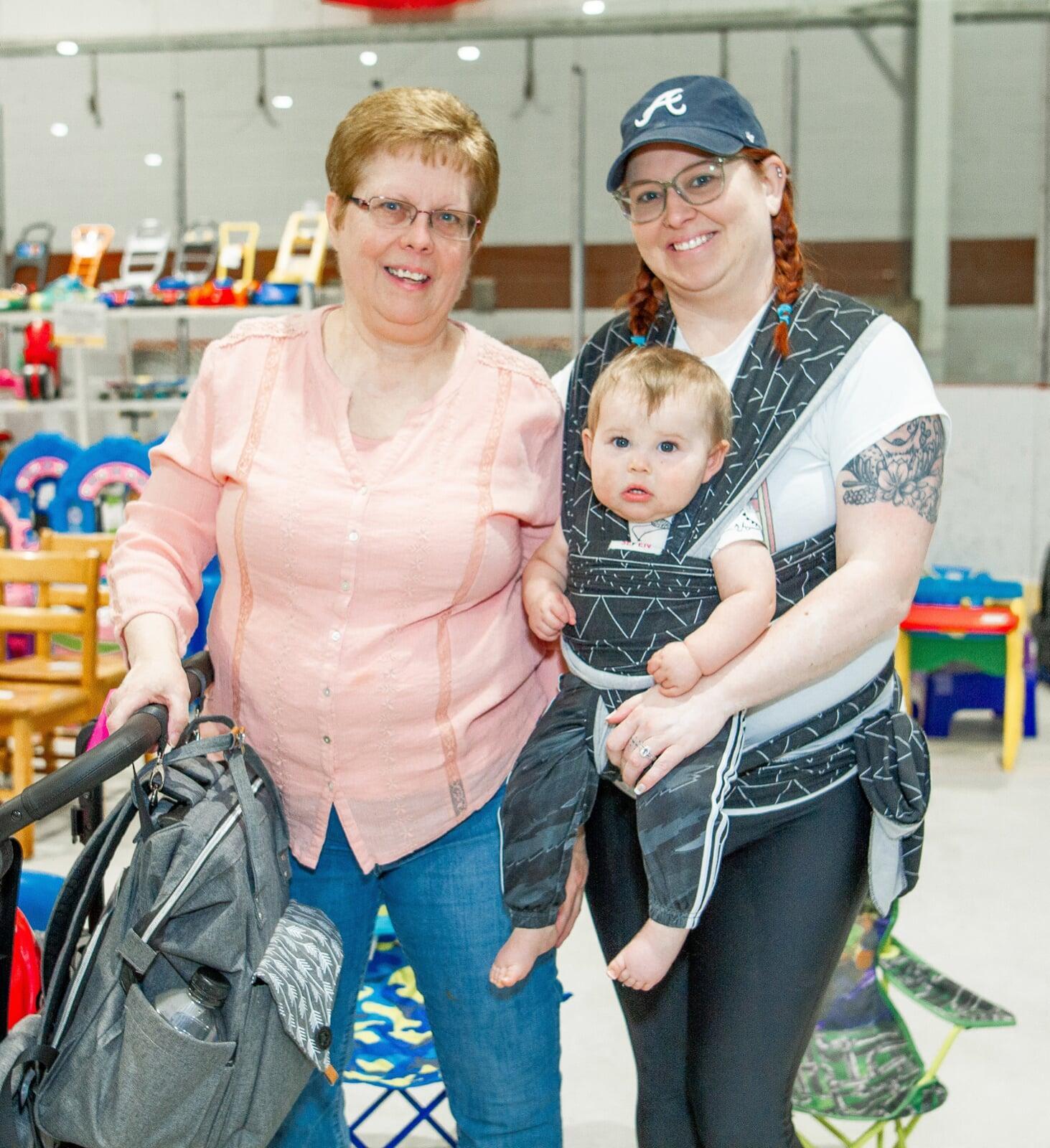 A mother carries her son in a front carrier. Grandmother is pushing the stroller. They are all shopping at the JBF sale and are surrounded by toys.