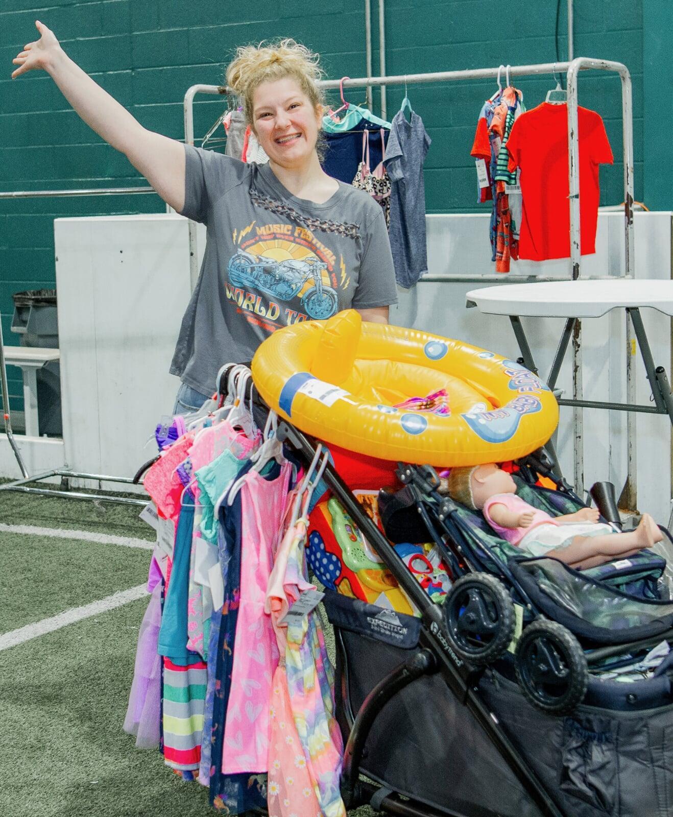 A woman waves with one hand and has the other on the handle of her wagon. The wagon is filled with toys and has girls clothing hanging from the handle.