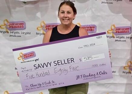 A woman holds up a pretend JBF consignor check (with her correct amount on it), and smiles.