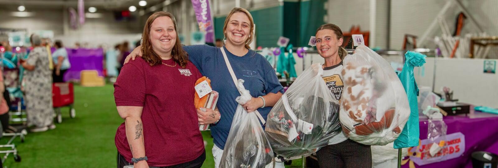 3 women stand in the register area holding items that they've just purchased at the Reading JBF sale.