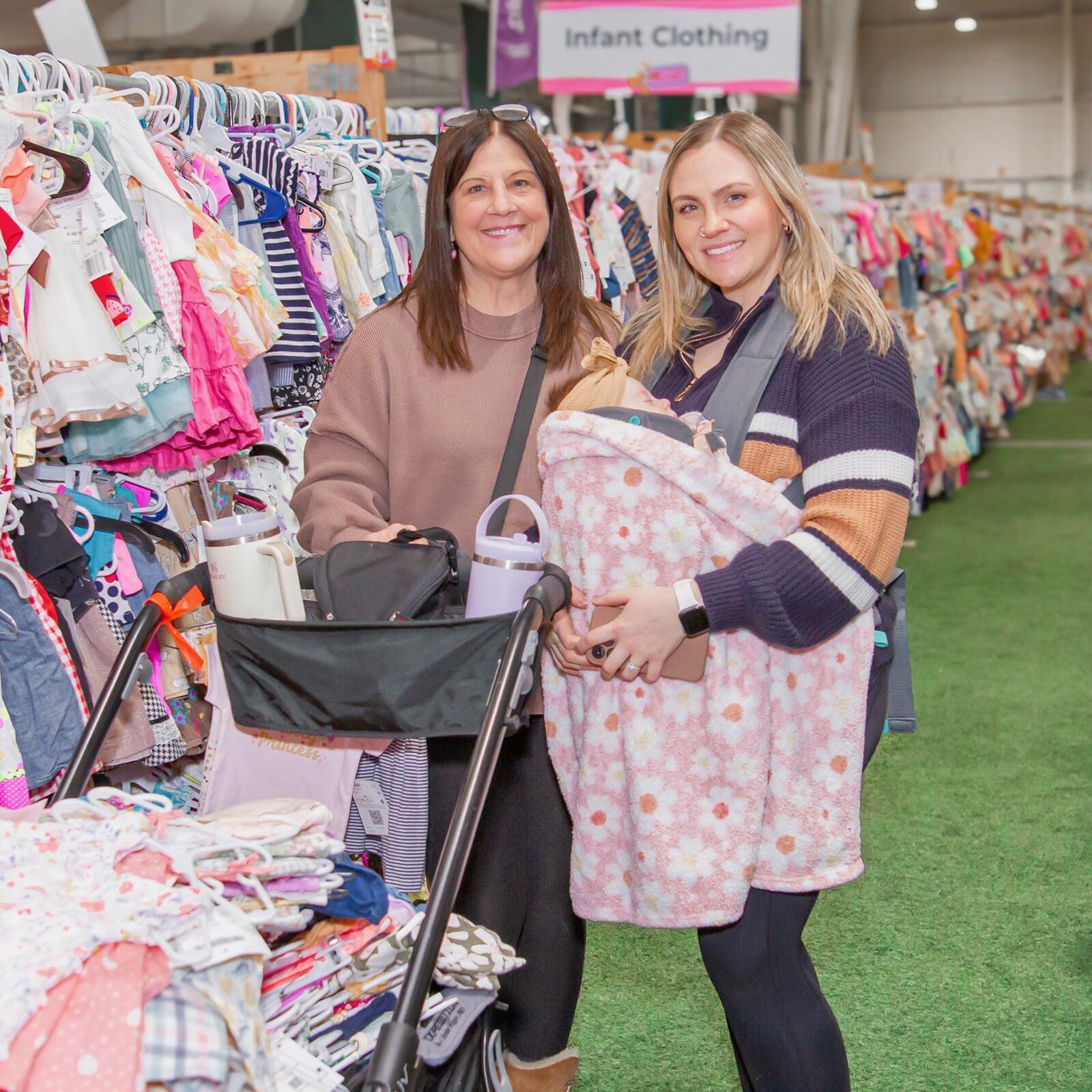 Two women shopping in the Infant Clothing section. One woman has a baby in a front carrier and the other woman is pushing a stroller.